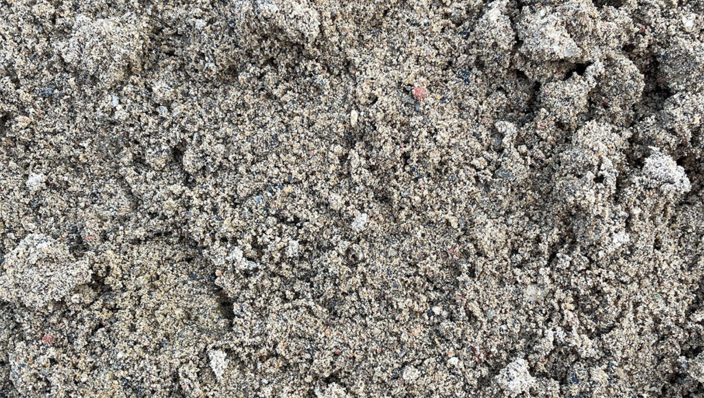 Close-up of loose, textured 0/2 recycled pipe bedding sand from Runfold, showing light brown and grey grains and small clumps.