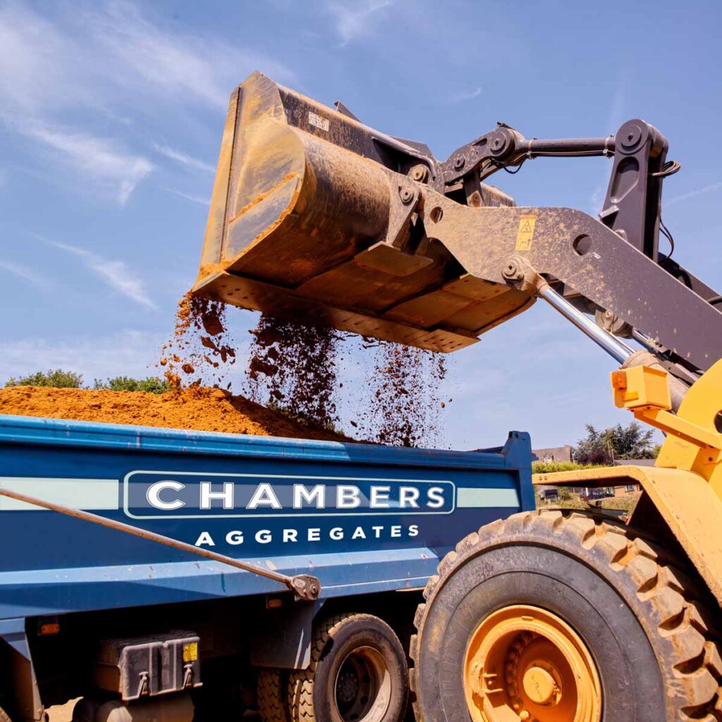 A construction vehicle pours soil into a blue "Chambers Aggregates" lorry at a site in Runfold, Farnham, Surrey.