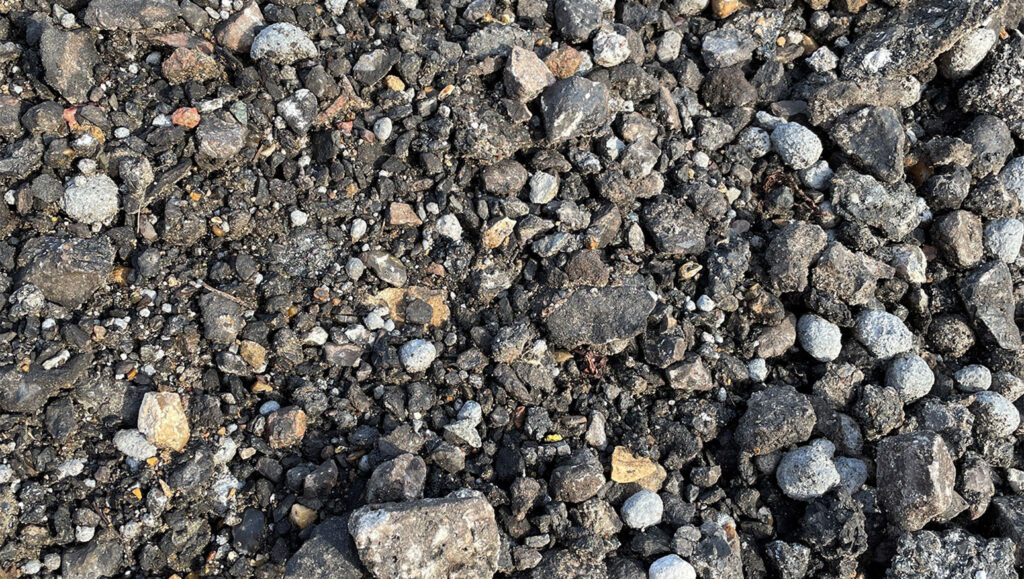 Close-up of rough asphalt road planings with uneven grey and brown stones, gravel, and debris in Runfold, Farnham, Surrey.