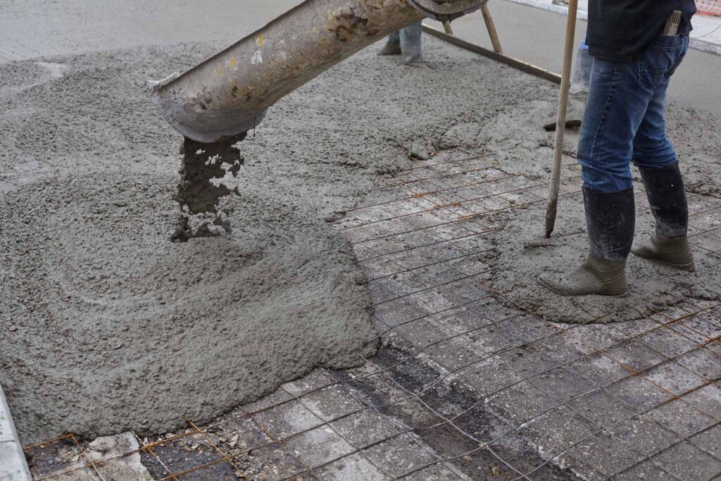 A person in boots spreads wet concrete from a chute over a rebar grid, preparing a foundation in Runfold, Farnham, Surrey.