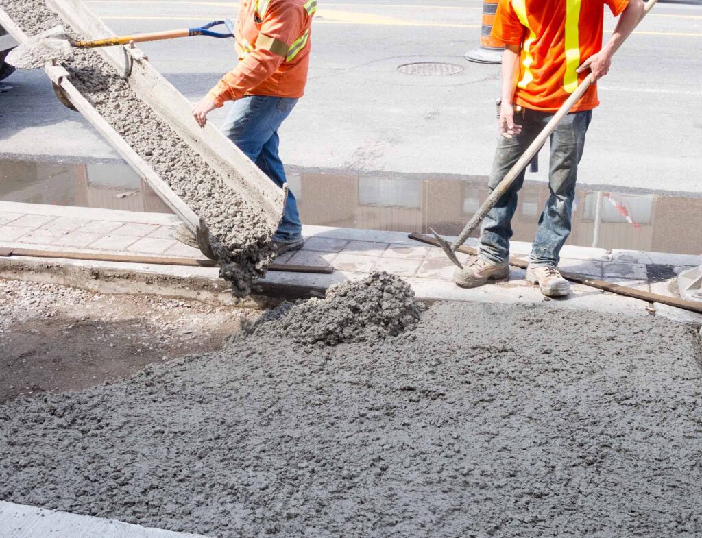 Two workers in orange shirts pour mixed on site concrete from a chute and spread it on a pavement repair in Runfold, Surrey.