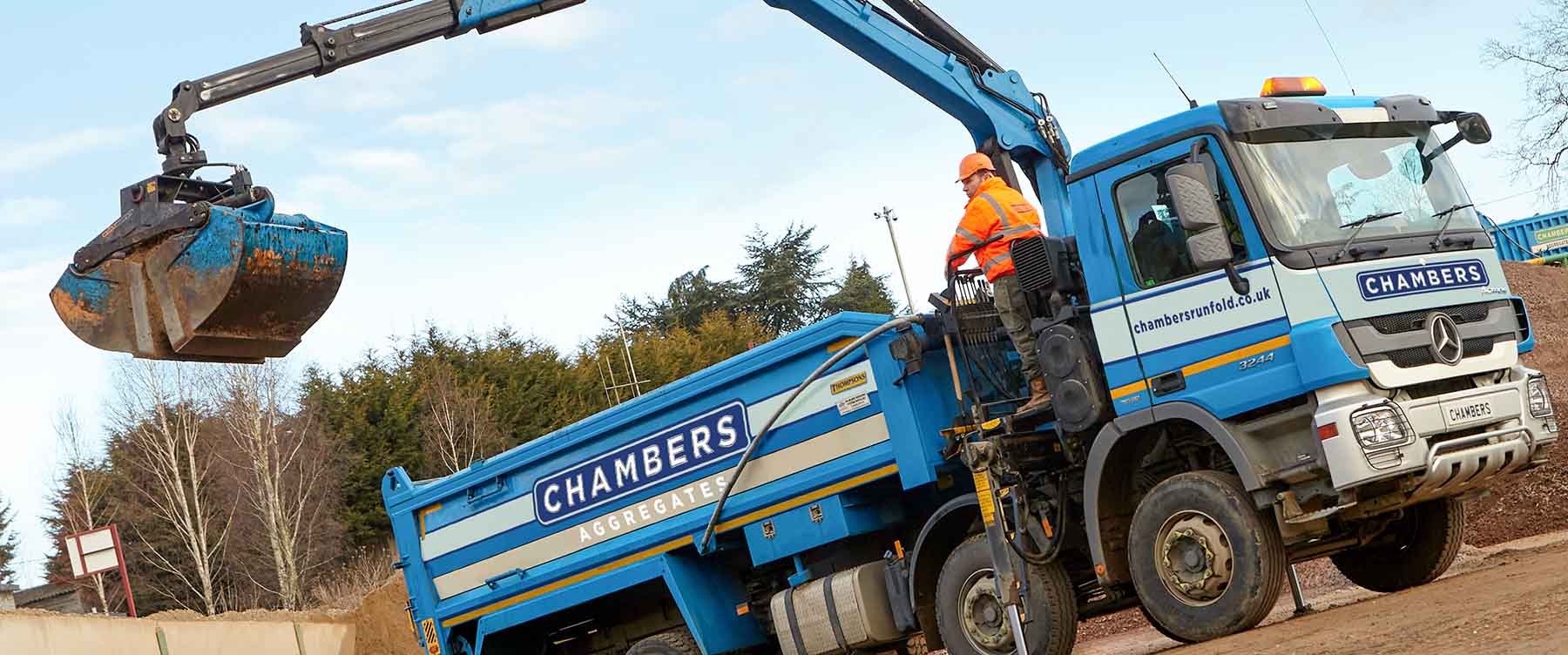 A worker uses a blue Chambers Aggregate lorry with crane and bucket to move soil at a Runfold, Surrey construction site.